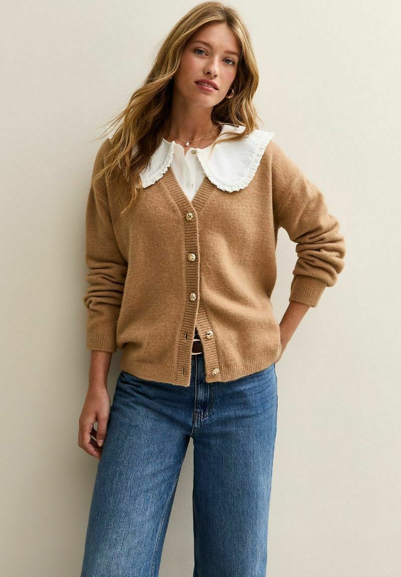 Bruine wollen cardigan met gouden knopen, gedragen over een witte blouse met een geplooide kraag, in combinatie met blauwe wijde jeans.