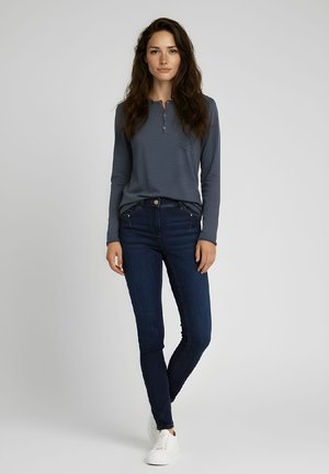 Mujer joven con cabello largo y castaño, vestida con una camiseta azul de manga larga, jeans ajustados oscuros y zapatillas blancas, de pie sobre un fondo gris.