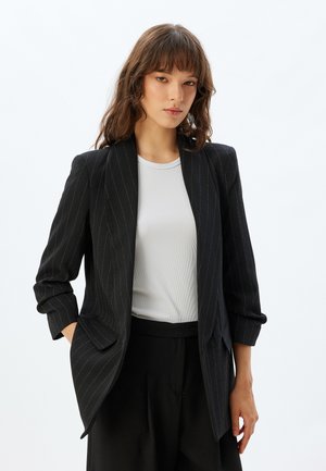 Blazer noir à rayures avec une coupe sur mesure, présentant un col châle et des poches latérales. Associé à une chemise blanche à côtes en dessous.