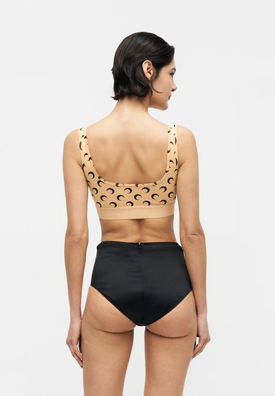 Γυναίκα με την πλάτη στραμμένη, φοράει μπεζ crop top με μαύρα σχέδια σε σχήμα ημισελήνου και ψηλόμεσο μαύρο εσώρουχο σε λευκό φόντο.