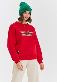 Czerwona bluza z napisem "mucho amor" w miętowym kolorze. Ściągacze przy mankietach i na dolnym końcu, miękka tkanina. W komplecie zielona dziana czapka.