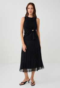Schwarzes ärmelloses Oberteil mit einem Drehdetail und goldenen Akzenten, kombiniert mit einem plissierten schwarzen Midirock und schwarzen Absatzsandalen.