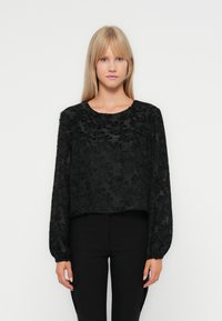 Svart langermet topp med blomstermønster, croppet design, rund halslinning og mykt stoff. Kombinert med svarte tettsittende bukser.