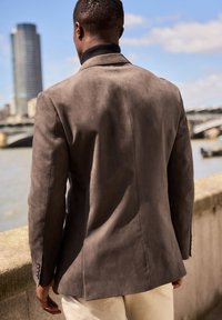 Blazer in suede marrone con una vestibilità su misura, dotato di rever a scollo, una fessura posteriore e chiusura a tre bottoni. Texture liscia, con dettagli minimi.