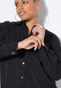 Camicia nera con bottoni, realizzata in un tessuto testurizzato. Presenta un colletto, maniche lunghe e bottoni lucidi. Accentuata da anelli argentati sulle mani.