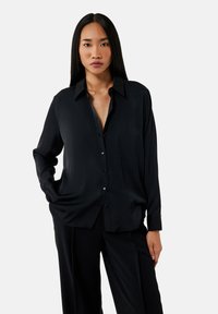 Camicia nera a maniche lunghe con bottoni, dotata di colletto e realizzata in un tessuto liscio. Presenta una vestibilità comoda e una leggera lucentezza. Abbinata a pantaloni neri.