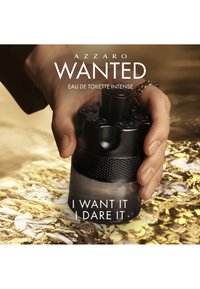 Hand hält eine schwarze Azzaro Wanted Eau de Toilette Intense Flasche über einer gold strukturierten Oberfläche mit sichtbarem Produktnamen und Slogan.