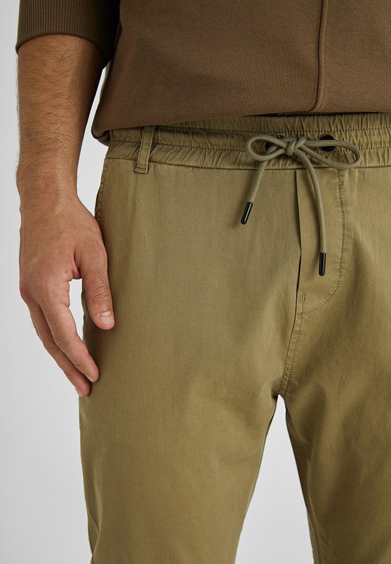 Helle olivgrüne Hose mit elastischer Taille und verstellbarem Kordelzug. Glattes Material, schmaler Schnitt, mit Seitentaschen und minimaler Naht.