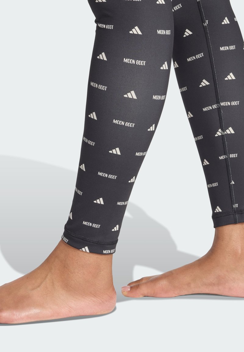 Leggings negros con un patrón de logotipo blanco repetido, que presentan un material suave y elástico y un ajuste ceñido en el tobillo, se muestran desde la pantorrilla hacia abajo.