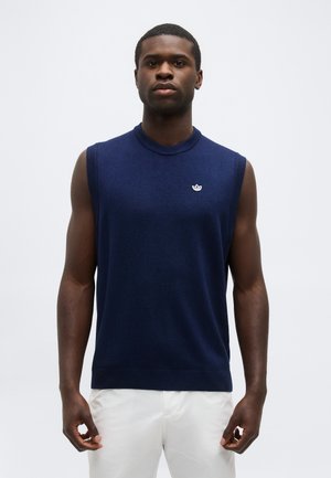 Άνδρας που φοράει αμάνικη πλεκτή μπλε σκούρα μπλούζα με ένα μικρό λευκό λογότυπο στο στήθος, συνδυασμένη με λευκό παντελόνι, στέκεται μπροστά σε απλό φόντο.