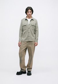 Νεαρός άνδρας ντυμένος με μπεζ πουκάμισο με κουμπιά πάνω από άσπρη μπλούζα, χαλαρούς καφέ τζιν και μαύρα χοντρά παπούτσια, μπροστά από λευκό φόντο.