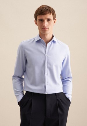 Camicia a maniche lunghe di colore blu chiaro con colletto button-down, caratterizzata da una leggerissima texture; indossata con pantaloni scuri a pieghe.