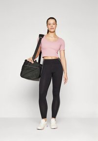 Fekete leggings oldalsávokkal, világos rózsaszín crop top és fehér sportcipő. Egy fekete texturált edzőtáskát visel.
