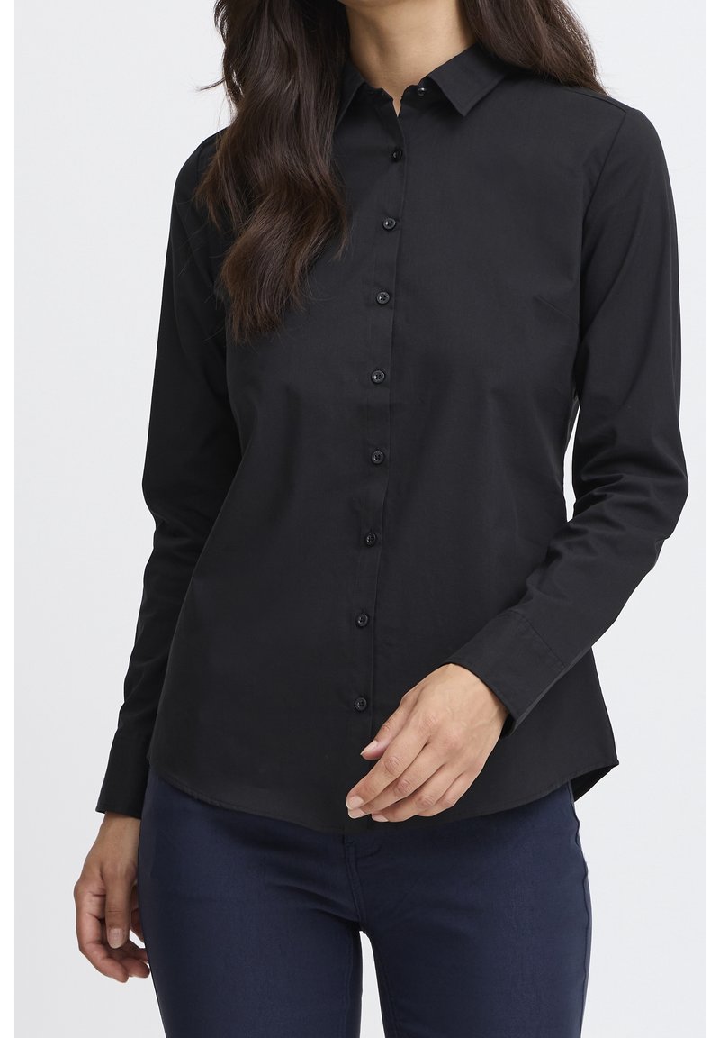 Femme portant une chemise noire ajustée boutonnée avec col et manches longues, associée à un pantalon bleu foncé, sur un fond uni.