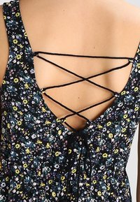 Musta kukkakuvioinen mekko, jossa syvä V-mallinen takakappale ristikkäisillä hihnoilla, ja kirkkaat keltaiset, violetit ja vihreät kuviot tummalla taustalla.