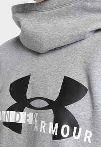 Grijze hoodie met een groot zwart Under Armour-logo en gedeeltelijk onleesbare witte tekst op de borst.