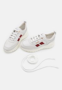 Valkotulit, joissa on punaiset sivuviivat, kultaiset "BALLY"-brändäykset, valkoiset nauhat ja ylimääräinen kierretty nauha valkoisella taustalla.