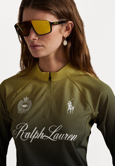 Gul og svart polo skjorte med prikket mønster, som har hvite logoer, en glidelås-krage og store svarte solbriller med speil-linser.