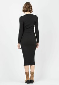 Robe noire en maille côtelée à manches longues et silhouette ajustée, s'évasant jusqu'à la mi-mollet. Associée à des bottines en daim marron.