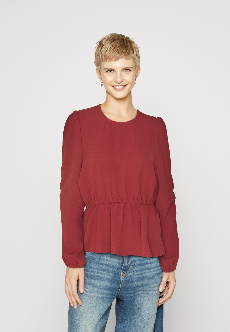 Haut peplum bordeaux à manches longues et décolleté rond, fabriqué en tissu texturé. Associé à un jean en denim bleu clair.