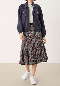 Veste de bombardier marine avec deux poches poitrine, jupe midi plissée à motif floral sombre, et baskets blanches.
