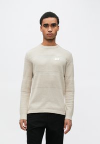 Beige strikkegenser med horisontale mønstre, rund hals og en liten etikett. Kombinert med svarte bukser.