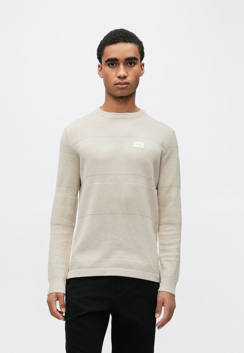 Beige strikkegenser med horisontale mønstre, rund hals og en liten etikett. Kombinert med svarte bukser.