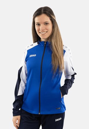 Giovane donna che indossa una giacca e pantaloni sportivi JAKO blu, bianca e nera, sorridente con una mano in tasca su uno sfondo semplice.
