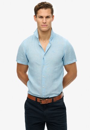 Chemise à manches courtes bleu clair en mélange de lin avec un col bien apparent, boutonnée sur le devant, et une texture subtile, portée avec un pantalon foncé et une ceinture marron.