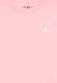 T-shirt in cotone rosa con colletto rotondo, caratterizzato dal logo Jumpman bianco sul lato sinistro del petto. Texture morbida e liscia, design minimale.