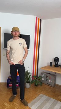 Camiseta de algodón beige con un parche de panda colorido, pantalones negros y zuecos marrones. El fondo presenta un televisor y rayas de colores en la pared.