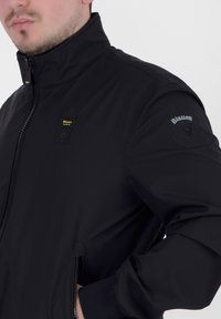 Giacca bomber nera con colletto alto, chiusura con zip e tasche con zip. Presenta un logo sulla manica e una texture liscia e opaca.