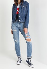 Woman wearing a denim jacket, white and red graphic t-shirt, ripped light blue jeans, and navy high-top sneakers against a white background.