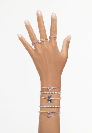 Mano con múltiples anillos: tres anchos en color rosa, dorado y transparente, junto con delicadas pulseras que presentan una flor, una luna y piedras preciosas.