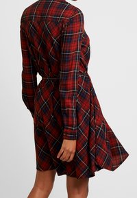 Punainen ja musta ruudullinen mekko pitkillä hihoilla, vyötäröltä kiristetty ja kellotettu helma. Sisältää nappikauluksen ja käänteiset hihansuut.