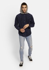 Camisa de botones azul marino con capucha gris, cordón y bolsillo frontal, combinada con jeans claros desgastados y zapatillas negras.