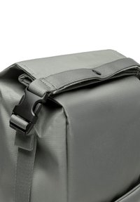 Sac à dos en tissu gris avec une surface texturée, sangle réglable et boucle en plastique pour une fermeture sécurisée. Forme compacte et rectangulaire.