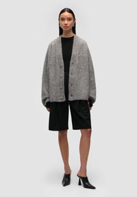 Szary sweter z dzianiny o prążkowanej fakturze, w oversize'owym fasonie, z pięcioma guzikami, noszony na czarnej bluzce i zestawiony z czarnymi szortami oraz szpilkami.