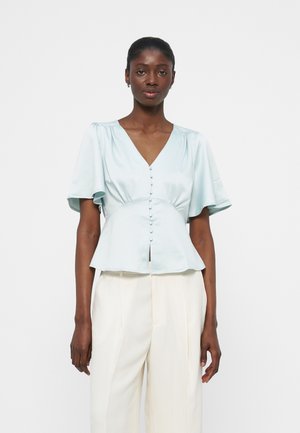 Tumeda nahaga naine, kes kannab hele sinist V-kaelusega nööbitavat pluusi lenduvate varrukatega ja kõrge vöökohaga valgeid pükse, seisab valge tausta ees.