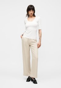 Vrouw met donker haar die een wit aansluitend topje draagt, beige wijde broekspijpen en zwarte loafers, staand met één hand in de zak.