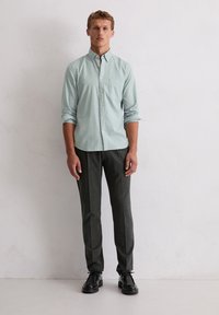 Camicia button-down azzurro chiaro con maniche lunghe, taschino sul petto, abbinata a pantaloni grigi scuri su misura e scarpe nere.