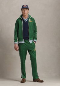 Trening verde cu dungi albe, compus dintr-o jachetă cu fermoar și pantaloni asortați. Purtați deasupra unui pulover tricotat bleumarin și a unei cămăși cu guler cu dungi.
