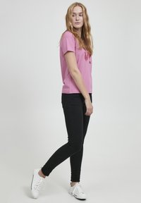 Lyserød bomulds T-shirt med korte ærmer, prydet med et blomsterdesign i rød. Bæres med sorte skinny jeans og hvide sneakers, på en neutral baggrund.