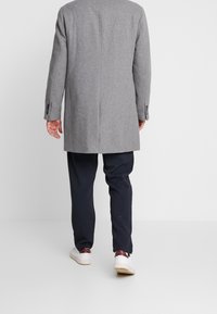 Cappotto di lana grigio chiaro con un design strutturato, mostrato da dietro. Abbinato a pantaloni scuri e scarpe da ginnastica bianche. Dettagli semplici e sobrii.