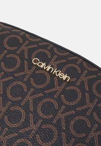 Schwarze strukturierte Oberfläche mit sich wiederholendem braunen "CK"-Muster und goldfarbenem "Calvin Klein"-Logo in der Mitte.
