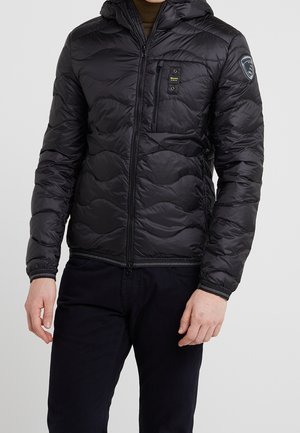 Mężczyzna ubrany w czarną pikowaną kurtkę na zamek błyskawiczny z kapturem, kieszeniami na piersi i bokach oraz naszywką na lewym rękawie.