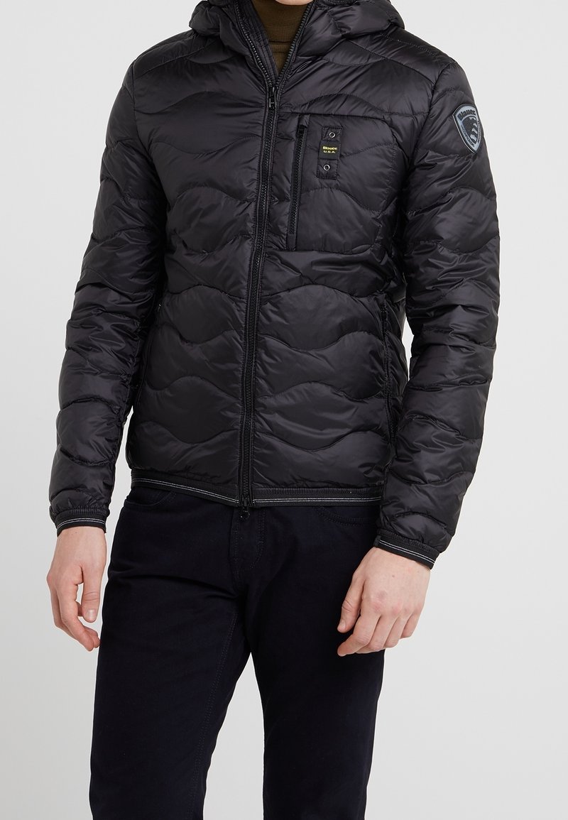 Homme portant une veste matelassée noire zippée avec capuche, poches poitrine et latérales, et un écusson sur la manche gauche.