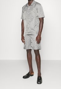 Grå kortärmad skjorta och matchande shorts i mjuk väv. Har en ficka, knappstängning och avslappnad passform. Svarta slip-on skor.