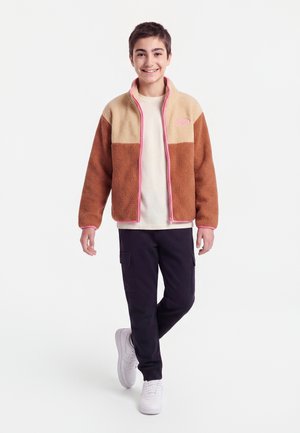 Lächelnde junge Person geht, trägt eine braune und beige Fleecejacke mit pinkem Besatz, weißes Hemd, schwarze Hose und weiße Sneakers vor weißem Hintergrund.