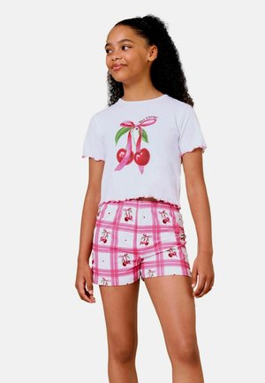 Mädchen mit lockigem Haar trägt ein weißes bauchfreies Top mit Kirschen und pink karierte Shorts, verziert mit Kirschmustern.